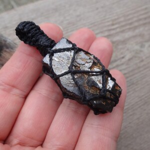 May include: A dark, irregularly shaped stone wrapped in a black woven cord. The stone has a metallic, silver-like sheen with areas of brown and black. The cord is tightly woven around the stone, creating a secure hold.