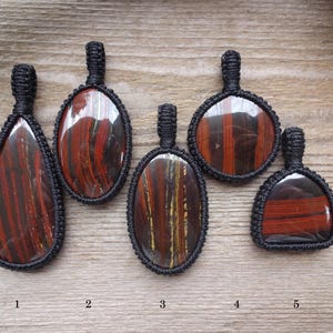 May include: Five handcrafted pendants with polished, striped red and brown stones, each encased in a black woven macrame setting. The pendants vary in shape, including teardrop and oval designs. The background is a light brown wooden surface.