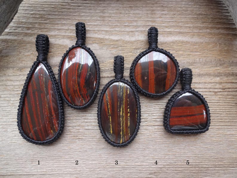 May include: Five handcrafted pendants with polished, striped red and brown stones, each encased in a black woven macrame setting. The pendants vary in shape, including teardrop and oval designs. The background is a light brown wooden surface.