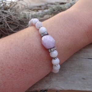 May include: A bracelet made with white and light purple beads. The bracelet has a larger, faceted, light purple bead with rhinestones.