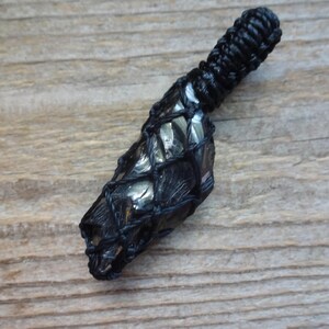 May include: A dark, metallic-looking stone pendant encased in a black macrame net. The stone has a faceted, reflective surface with deep gray and charcoal tones. The top of the pendant features a tightly knotted black cord loop for suspension.