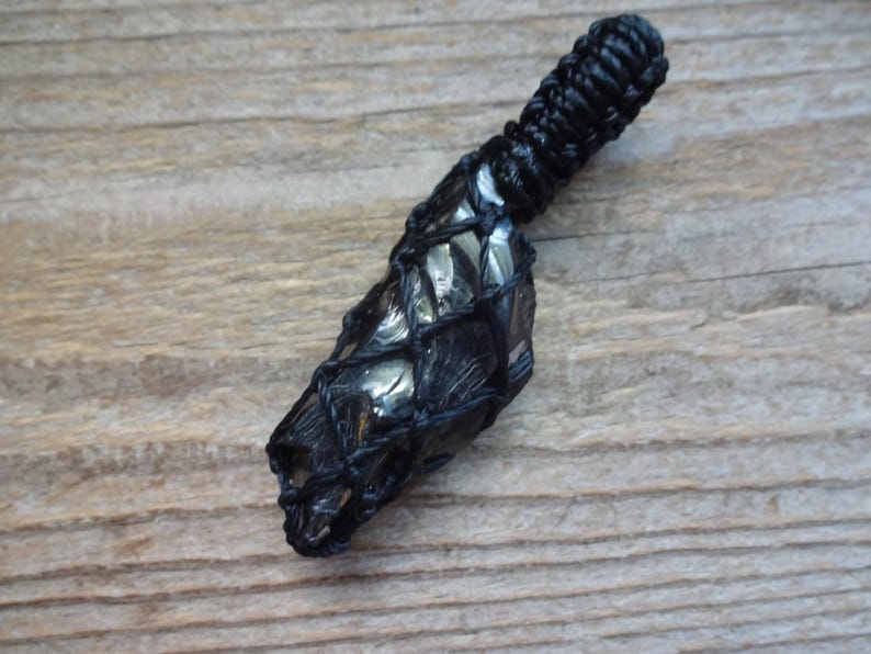 May include: A dark, metallic-looking stone pendant encased in a black macrame net. The stone has a faceted, reflective surface with deep gray and charcoal tones. The top of the pendant features a tightly knotted black cord loop for suspension.