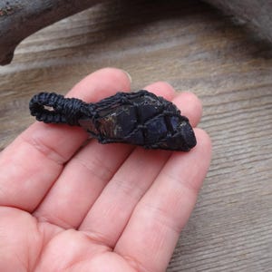 May include: A dark blue crystal pendant wrapped in black macrame cord. The crystal has a rough, natural texture and is held in a woven net, creating a unique and earthy aesthetic. The pendant is shown against a wooden background.
