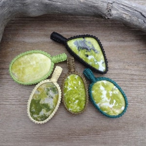 May include: Five green and white gemstone pendants with knotted cord wrappings in various colors, including black, green, and cream. The pendants are arranged on a wooden surface.