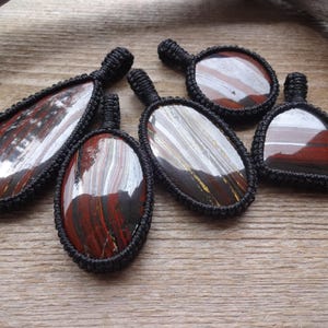 May include: Six gemstone pendants with a black woven cord border. The stones have a mix of red, brown, white, and yellow hues. The pendants are various shapes, including oval and teardrop, and are displayed on a wooden surface.