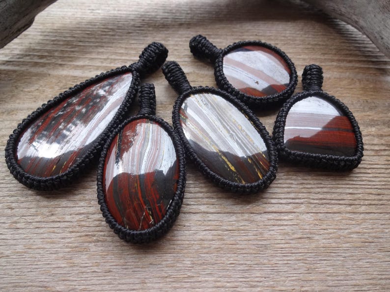 May include: Six gemstone pendants with a black woven cord border. The stones have a mix of red, brown, white, and yellow hues. The pendants are various shapes, including oval and teardrop, and are displayed on a wooden surface.