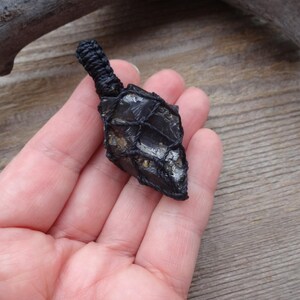 May include: A black obsidian arrowhead pendant, intricately wrapped in black cord. The arrowhead is held in a hand, showcasing its dark, polished surface and unique shape. The pendant's design suggests a focus on natural materials and craftsmanship.