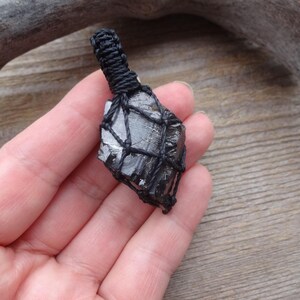 May include: A black crystal pendant wrapped in a black woven net. The crystal is a dark gray color with a rough texture. The pendant is held in a hand against a wooden background.