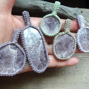 May include: Five pendants featuring translucent, irregular purple stone slices encased in knotted cord frames. The frames are colored in shades of light purple, sage green, and beige, each with a small loop at the top for hanging as a necklace.