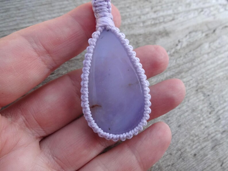 Peut inclure: Pendentif en forme de goutte d'eau avec une pierre violet clair, entour&eacute; d'un motif macram&eacute;. Le pendentif est suspendu &agrave; un cordon assorti. La pierre a une surface lisse et est de couleur lavande.
