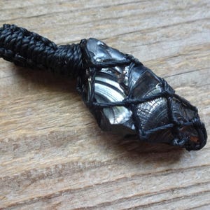 May include: A dark, glossy, irregular-shaped stone pendant encased in a black knotted cord netting. The stone features a reflective, metallic-like surface with visible conchoidal fractures. A thick, braided black cord loop extends from the top.