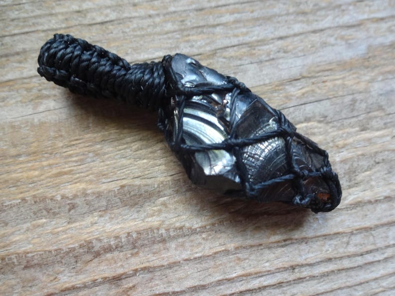 May include: A dark, glossy, irregular-shaped stone pendant encased in a black knotted cord netting. The stone features a reflective, metallic-like surface with visible conchoidal fractures. A thick, braided black cord loop extends from the top.