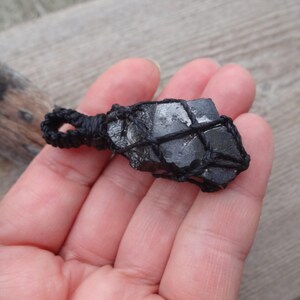 May include: A black crystal pendant, held in a hand, is encased in a woven black net. The crystal has a rough, natural texture and a dark, reflective surface. The pendant is suspended from a loop of black cord, suggesting it's a necklace.