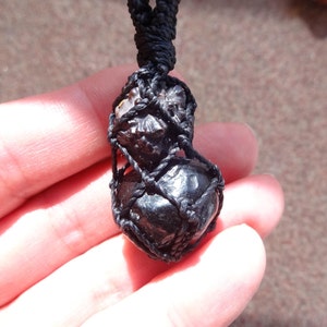 May include: Black obsidian gemstone pendant wrapped in black macrame cord. The pendant is round and has a shiny, polished finish.