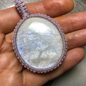 May include: A white moonstone pendant with a lavender cord. The moonstone is oval shaped and has a smooth, polished surface. The cord is tied in a knot at the top of the pendant.