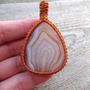 May include: A brown cord wrapped around a white and brown agate pendant. The pendant is teardrop shaped and has a natural, organic look.