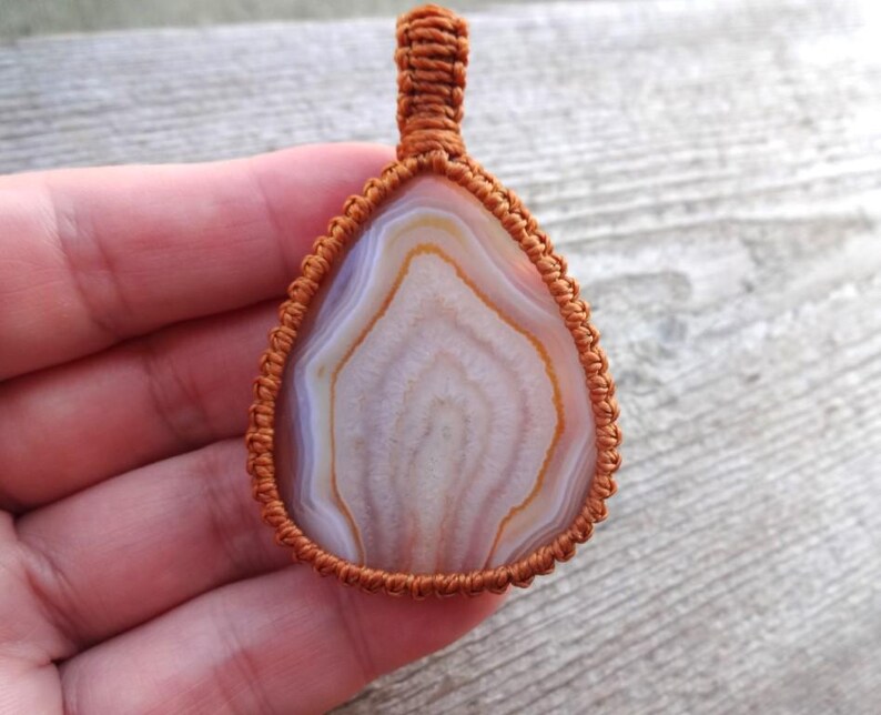May include: A brown cord wrapped around a white and brown agate pendant. The pendant is teardrop shaped and has a natural, organic look.