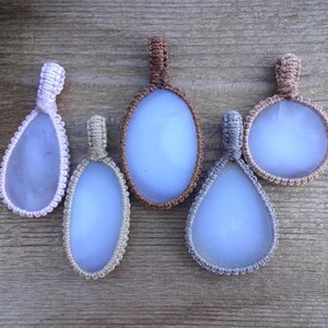 Peut inclure: Cinq pendentifs macram&eacute; faits &agrave; la main avec des pierres bleu clair. Les pendentifs ont diff&eacute;rentes formes, dont ovales et en forme de goutte, et sont encadr&eacute;s de cordons macram&eacute; de diff&eacute;rentes couleurs, comme le rose, le beige, le marron et le gris. Les pendentifs sont expos&eacute;s sur une surface en bois.