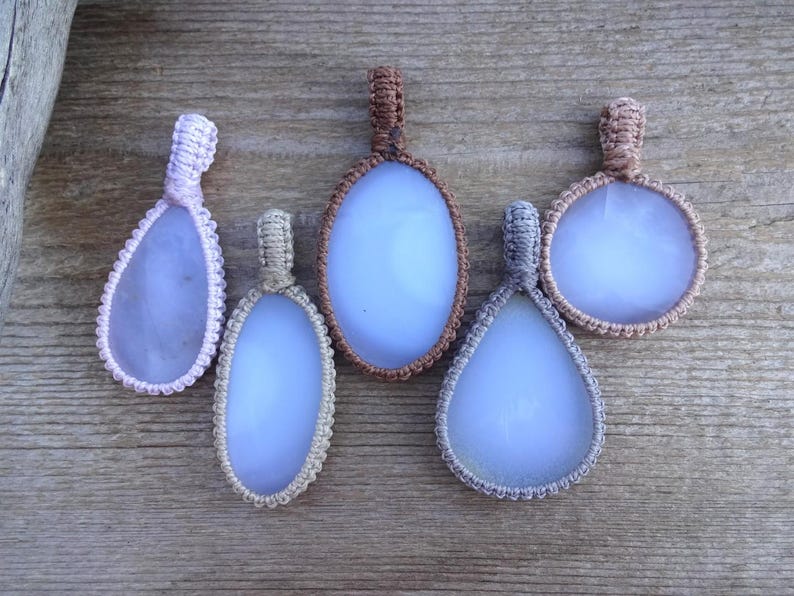 Peut inclure: Cinq pendentifs macram&eacute; faits &agrave; la main avec des pierres bleu clair. Les pendentifs ont diff&eacute;rentes formes, dont ovales et en forme de goutte, et sont encadr&eacute;s de cordons macram&eacute; de diff&eacute;rentes couleurs, comme le rose, le beige, le marron et le gris. Les pendentifs sont expos&eacute;s sur une surface en bois.