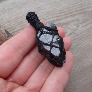May include: A black obsidian crystal pendant wrapped in a black woven net. The crystal is a dark, glossy color and is held in a macrame-style wrap. The pendant is held in a hand against a wooden background.