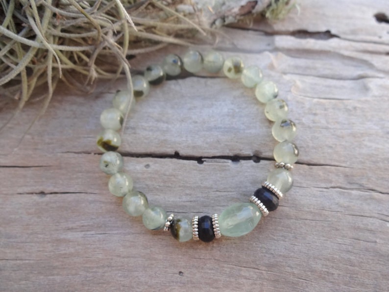 May include: A green and black gemstone bracelet with silver accents. The bracelet is made of round beads and a larger oval bead.