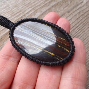 May include: An oval-shaped pendant featuring a polished stone with brown, yellow, and gray striations, encased in a black woven macrame setting. The pendant has a small loop at the top for hanging.