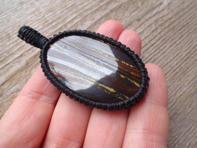 May include: An oval-shaped pendant featuring a polished stone with brown, yellow, and gray striations, encased in a black woven macrame setting. The pendant has a small loop at the top for hanging.