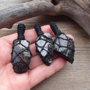 May include: Three raw black tourmaline pendants, each encased in a black woven net. The pendants are displayed against a natural wood background, showcasing their dark, textured surfaces and unique shapes. The pendants are approximately 1.5 inches in length.