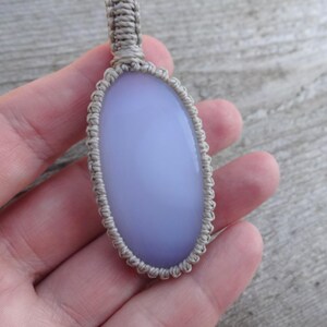 Peut inclure: Un pendentif ovale bleu clair avec un cordon en macram&eacute;. Le pendentif est une pierre lisse et polie, et le cordon en macram&eacute; est de couleur beige neutre. Le pendentif est tenu dans une main.