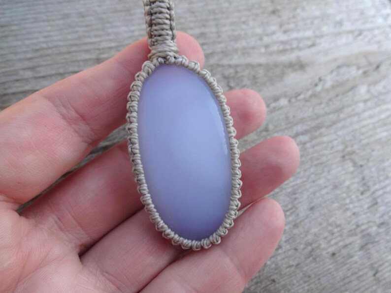 Peut inclure: Un pendentif ovale bleu clair avec un cordon en macram&eacute;. Le pendentif est une pierre lisse et polie, et le cordon en macram&eacute; est de couleur beige neutre. Le pendentif est tenu dans une main.