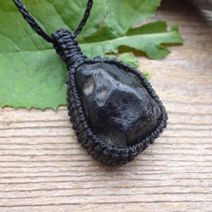May include: A black stone pendant with a woven black cord. The stone is irregularly shaped and set within a woven black frame. The pendant is displayed on a wooden surface with a green leaf.