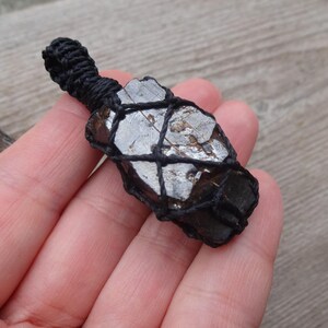 May include: A dark, irregularly shaped stone pendant wrapped in black macrame. The stone has a metallic, reflective surface and is suspended from a woven loop. The pendant is held in a hand against a wooden background.