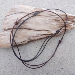 May include: Two leather cords, one brown and one black, are tied together with knots. The cords are lying on a piece of driftwood.