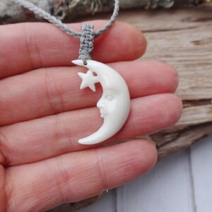 May include: A white bone pendant carved in the shape of a crescent moon with a star. The moon has a face carved into it.