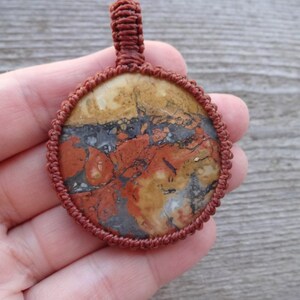 May include: A round, red, brown, and white stone pendant with a knotted cord border. The pendant is held in a hand.