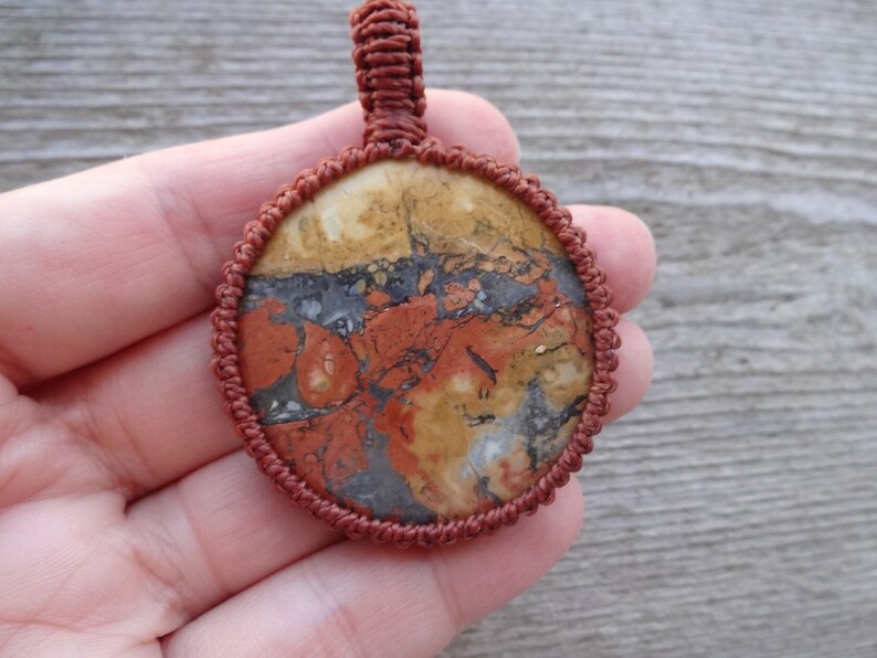 May include: A round, red, brown, and white stone pendant with a knotted cord border. The pendant is held in a hand.