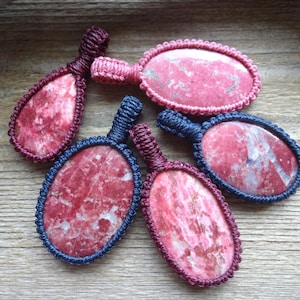 May include: Five pendants featuring polished pink and white mottled stones encased in intricate macrame cord frames. The collection includes one teardrop-shaped pendant and four oval-shaped pendants, each with a matching cord loop for hanging.