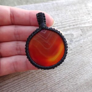 May include: A red agate pendant with black macrame cord. The agate has a swirling pattern of red, orange, and yellow.
