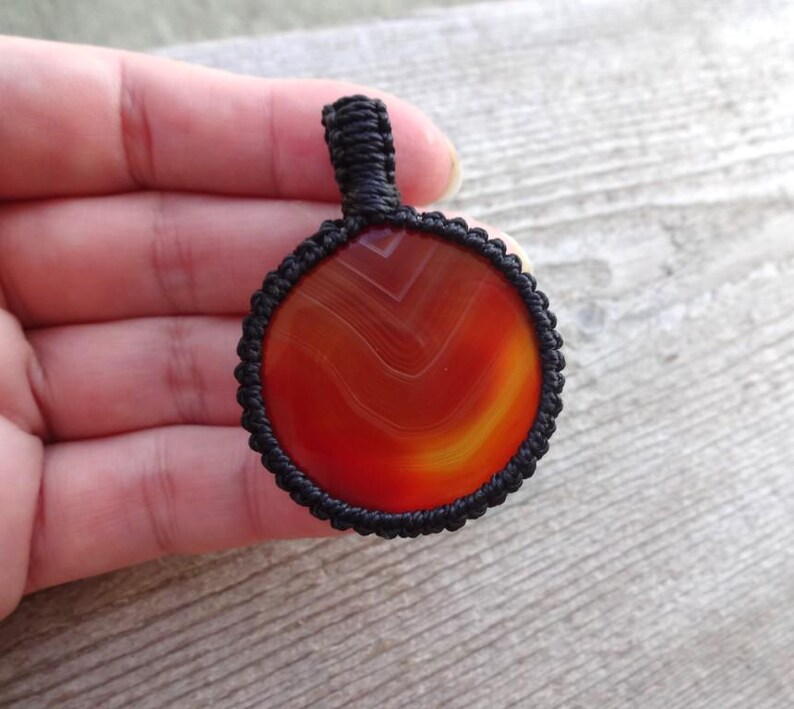 May include: A red agate pendant with black macrame cord. The agate has a swirling pattern of red, orange, and yellow.