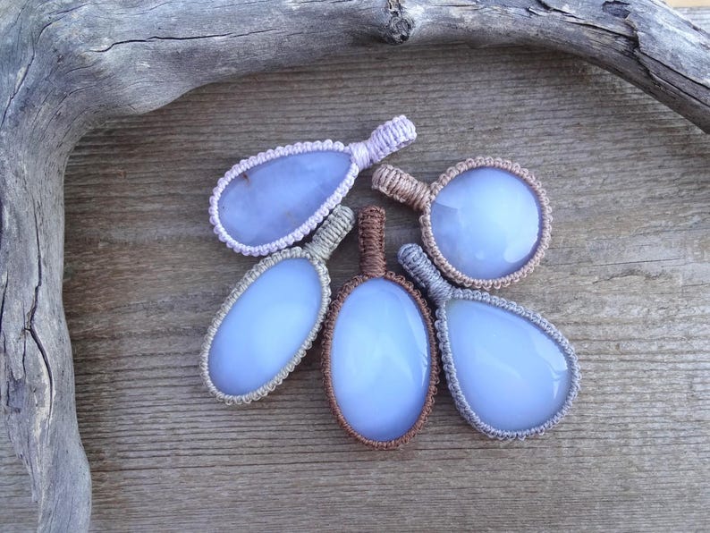 Peut inclure: Cinq pendentifs en calc&eacute;doine bleue avec des d&eacute;tails en macram&eacute; de diff&eacute;rentes formes : ovale, goutte d'eau et rond. Les pendentifs sont pos&eacute;s sur un fond en bois rustique, avec un morceau de bois flott&eacute; dans le coin sup&eacute;rieur gauche.