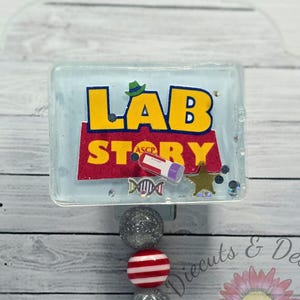 May include: A badge reel with a rectangular design featuring the words "LAB STORY" in yellow and red, with a green hat and other graphics. The reel has three beads: two silver glitter and one red and white striped. A silver clip is attached.