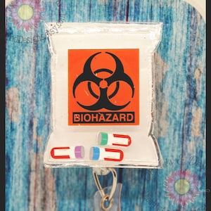 May include: A clear plastic badge holder with a biohazard symbol and the word "BIOHAZARD" printed on it. The holder has three colorful pill-shaped charms attached to it.