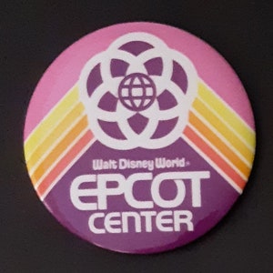 May include: A purple and pink button with a white logo that says "Walt Disney World - EPCOT CENTER". The logo features a purple sphere with a white circle around it and a white starburst design.