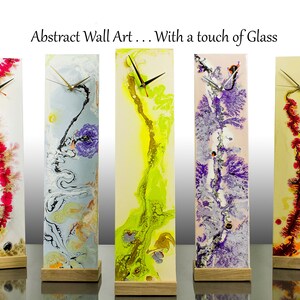 Op de afbeelding: Vijf abstracte wandklokken met verschillende kleurrijke ontwerpen op glas. Elke klok heeft een houten voet en een zilveren wijzerplaat met zwarte wijzers.