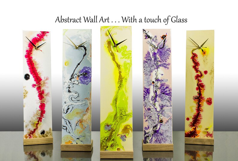 Op de afbeelding: Vijf abstracte wandklokken met verschillende kleurrijke ontwerpen op glas. Elke klok heeft een houten voet en een zilveren wijzerplaat met zwarte wijzers.