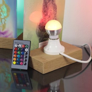 Op de afbeelding: Een witte gloeilamp met een afstandsbediening om de kleur van het licht te veranderen. De gloeilamp staat op een houten voet en is verbonden met een wit snoer. De afstandsbediening heeft een verscheidenheid aan kleurknoppen, evenals knoppen voor wit licht en een knipperende modus.