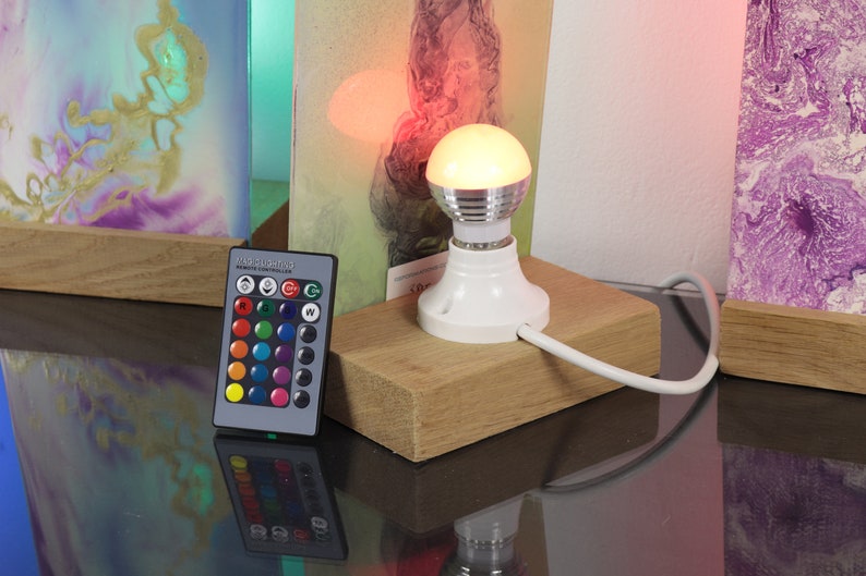 Op de afbeelding: Een witte gloeilamp met een afstandsbediening om de kleur van het licht te veranderen. De gloeilamp staat op een houten voet en is verbonden met een wit snoer. De afstandsbediening heeft een verscheidenheid aan kleurknoppen, evenals knoppen voor wit licht en een knipperende modus.