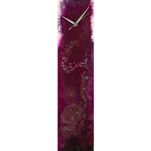 Pode incluir: Um relógio de parede vertical com um mostrador e ponteiros prateados. O mostrador do relógio está definido contra um fundo roxo escuro com manchas prateadas.