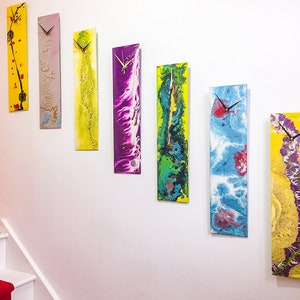 Op de afbeelding: Zeven kleurrijke abstracte klokken met zwarte wijzers aan een witte muur. De klokken zijn in verschillende kleuren, waaronder geel, paars, blauw en groen.