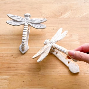 May include: Two white cast iron dragonfly door knockers. The knockers are in the shape of a dragonfly with a long, slender body and delicate wings. The knockers are mounted on a wooden surface.